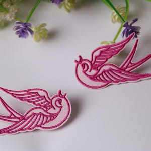 Może przedstawiać: Dwa różowe haftowane naszywki z latającymi jaskółkami.