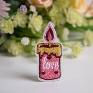 Può includere: Toppa ricamata a forma di candela rosa con la parte superiore dorata e una fiamma rossa. La parola "love" è ricamata in bianco sulla candela, con due piccoli dettagli a forma di cuore. La toppa è su una superficie bianca con fiori sullo sfondo.