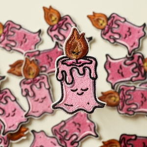 Può includere: Una collezione di toppe da stiro ricamate rosa che presentano una candela carina con un viso sorridente e una fiamma. La candela sta gocciolando cera e ha un design semplice.
