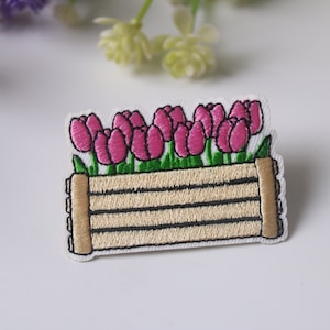 Op de afbeelding: Geborduurde strijk-patch met een houten plantenbak gevuld met roze tulpen. De plantenbak is lichtbruin met donkerbruine lijnen.