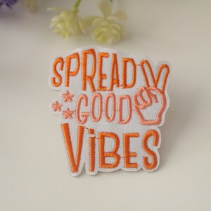 May include: White embroidered patch with the text "Spread Good Vibes" in orange and pink thread. A peace sign is included in the design.