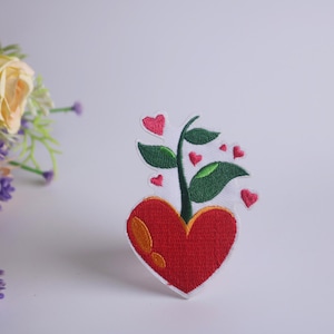 以下が含まれることがあります： 赤いハートに緑の植物が生えている刺繍のアイロン接着パッチ。植物には小さなピンクのハートが枝から生えています。