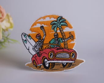 May include: Embroidered patch featuring two skeletons in a red car driving on a beach with a surfboard and palm tree. The background is a sunset with clouds.