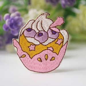 May include: A pink and yellow embroidered patch with a white frosting design and purple berries. The patch has a star and moon design and is perfect for adding a whimsical touch to clothing or accessories.