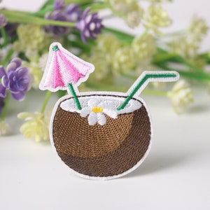Pode incluir: Etiqueta bordada de um coco castanho com uma flor branca, um canudo verde e um guarda-chuva rosa.