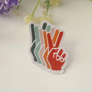 Puede incluir: Un parche de signo de paz multicolor con un contorno blanco. El parche está hecho de fieltro y tiene un diseño en capas con tonos de verde, naranja y rojo.