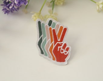 Può includere: Una toppa a forma di segno della pace multicolore con un contorno bianco. La toppa è realizzata in feltro e presenta un design a strati con sfumature di verde, arancione e rosso.