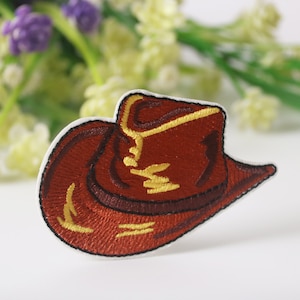 May include: A brown cowboy hat embroidered patch with yellow stitching. The patch is on a white background.