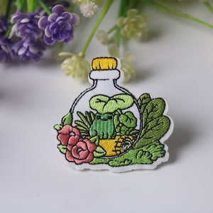 Peut inclure: Patch brodée thermocollante représentant une grenouille verte à l'intérieur d'une bouteille en verre avec un bouchon jaune. La bouteille est remplie de feuilles vertes, d'une fleur rose et d'une abeille jaune.