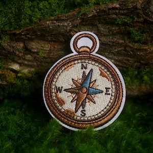 May include: A circular embroidered patch featuring a compass design with a brown border, a cream-colored face, and a central star-shaped needle in blue and brown. The letters N, E, S, and W are stitched in black around the compass dial.