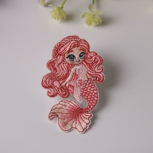 Può includere: Toppa ricamata di una sirena con capelli e coda rosa. La sirena ha gli occhi blu e un'espressione sorridente. La toppa è bordata di bianco ed è destinata all'abbigliamento o agli accessori.