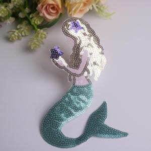 May include: A sequined mermaid patch in shades of teal, silver, and lavender. The mermaid holds a purple starfish. The patch has a teal tail, a silver sequined body, and silver hair. The mermaid is a decorative item.
