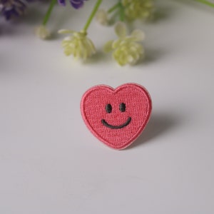 May include: A pink heart-shaped patch with a black embroidered smiley face.