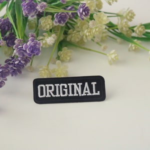 May include: A black fabric patch with white embroidery that reads "ORIGINAL". The patch is surrounded by purple and white flowers.