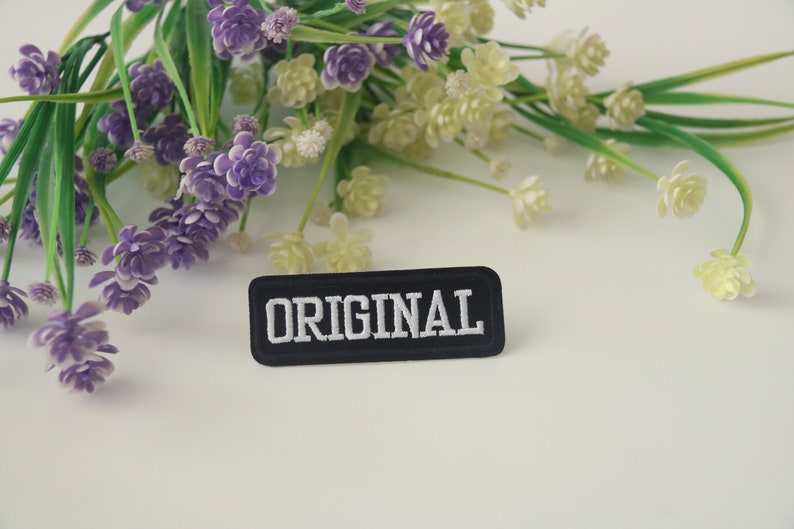 May include: A black fabric patch with white embroidery that reads "ORIGINAL". The patch is surrounded by purple and white flowers.