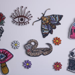 May include: A set of nine embroidered iron-on patches featuring various designs, including a rose, an eye, a moth, a skull with a candle, a snake, a mushroom with an eye, and three daisies.