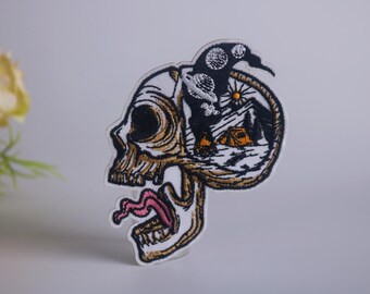 May include: White embroidered patch featuring a skull with a camping scene inside. The skull has a pink tongue sticking out. The camping scene includes a tent, mountains, a sun, and clouds.