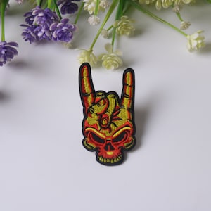 May include: An embroidered iron-on patch featuring a yellow and red skull with a hand making the rock and roll hand gesture.