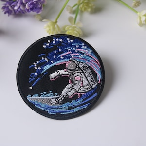 May include: Embroidered patch featuring an astronaut riding a surfboard on a wave of blue and pink with white stars on a black background.
