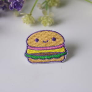 Pode incluir: Remendo bordado em forma de hambúrguer sorridente. O remendo apresenta um pão castanho claro, recheios rosa, amarelos e verdes e um contorno roxo. O remendo está sobre uma superfície branca com flores roxas no fundo.