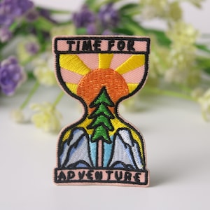 May include: An embroidered patch with a pink background and the text "TIME FOR ADVENTURE". The patch features an hourglass with a sun setting in the top and a green pine tree in the bottom.