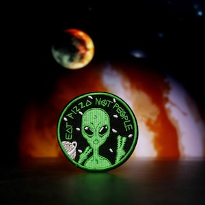 Puede incluir: Parche bordado de un extraterrestre verde con el texto "Eat Pizza Not People" sobre un fondo negro. El parche está sobre una superficie oscura con un fondo borroso naranja y blanco.