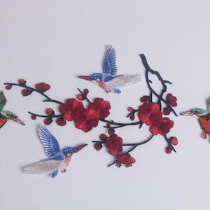Può includere: Toppe ricamate raffiguranti uccelli colorati e fiori rossi su rami neri. Gli uccelli hanno ali blu, arancioni, verdi e gialle. I fiori sono di un colore rosso intenso. Le toppe sono probabilmente per abbigliamento o progetti artigianali.