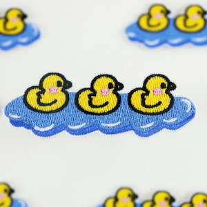 May include: Three yellow rubber duckies with black eyes and pink beaks are floating on a blue cloud-shaped patch. The duckies are embroidered on a white fabric.