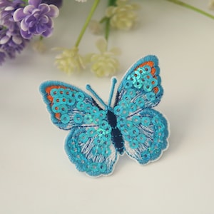 Puede incluir: Un parche bordado en forma de mariposa azul con lentejuelas. La mariposa tiene detalles naranjas en las alas.