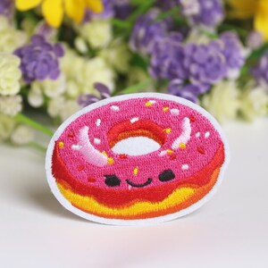 Peut inclure: Patch brodé en forme de donut rose avec glaçage jaune et pépites blanches. Le donut a un visage souriant avec des yeux noirs et un sourire.