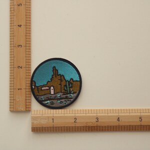 Peut inclure: &Eacute;cusson rond brod&eacute; repr&eacute;sentant un paysage d&eacute;sertique avec un ciel turquoise. L'&eacute;cusson repr&eacute;sente des formations rocheuses brunes, un cactus et un b&acirc;timent avec une porte rose. Le patch mesure environ 5 cm de diam&egrave;tre.