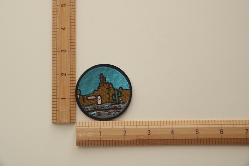 Peut inclure: &Eacute;cusson rond brod&eacute; repr&eacute;sentant un paysage d&eacute;sertique avec un ciel turquoise. L'&eacute;cusson repr&eacute;sente des formations rocheuses brunes, un cactus et un b&acirc;timent avec une porte rose. Le patch mesure environ 5 cm de diam&egrave;tre.