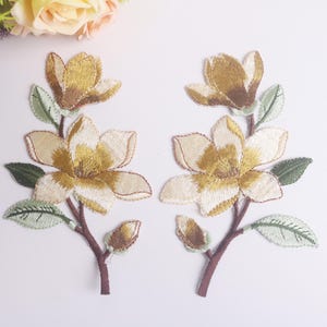 Pode incluir: Dois patches florais bordados com flores de magnólia creme e douradas com folhas verdes e caules castanhos. Estes patches são ideais para adicionar um toque decorativo a roupas ou acessórios.