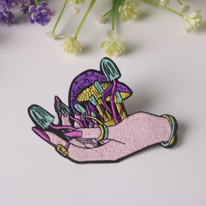 Könnte beinhalten: Ein gestickter Aufnäher mit einer Hand, die einen Haufen lila, grüner und gelber Pilze hält. Die Hand ist rosa und hat goldene Ringe an den Fingern.
