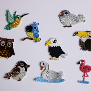 Könnte beinhalten: Eine Sammlung gestickter Vogelaufnäher, darunter ein Kolibri, ein Papagei, eine Eule, ein Adler, ein Tukan, ein Spatz, ein Schwan und ein Flamingo. Jeder Aufnäher hat detaillierte Stickereien und leuchtende Farben.