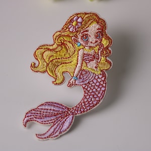 Può includere: Toppa ricamata raffigurante una sirena dei cartoni animati con lunghi capelli biondi, una coda rosa e un fiore tra i capelli. La sirena indossa un top a conchiglia rosa e un braccialetto blu. La toppa è progettata con cuciture dettagliate.