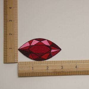 May include: An embroidered gem patch in red and pink. The gem is teardrop shaped and has a faceted design.