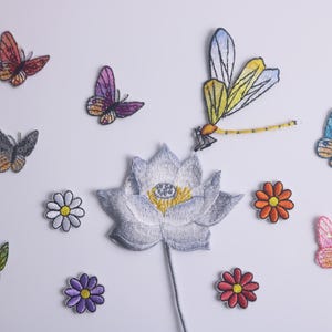May include: A collection of embroidered patches depicting butterflies, flowers, and a dragonfly. The patches include a white lotus flower, daisies, and butterflies in shades of red, purple, blue, and green. These patches are suitable for embellishing clothing or accessories.
