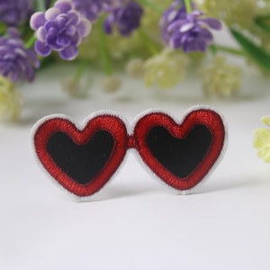Puede incluir: Gafas de sol de corazón rojo con lentes negros. Las gafas están bordadas en una tela blanca.