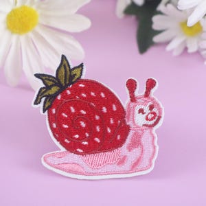 May include: Embroidered patch featuring a whimsical strawberry snail design. The snail's shell is a vibrant red strawberry with white seeds, and the body is pink. The patch is outlined in white, with a light purple background and daisies.
