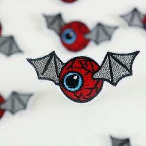 Peut inclure: Œil rouge avec un iris et une pupille bleus, avec des ailes de chauve-souris grises de chaque côté. Le globe oculaire est brodé sur un tissu blanc.