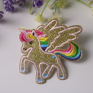 May include: A gold glitter unicorn with rainbow mane and tail, pink wings, and blue hooves. The unicorn is embroidered with glitter and thread.