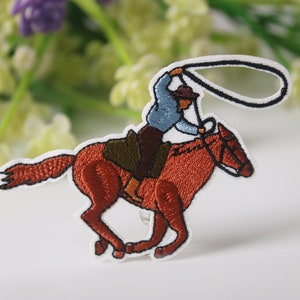 May include: Embroidered iron-on patch featuring a cowboy riding a brown horse and holding a lasso. The cowboy is wearing a blue shirt, brown pants, and a brown hat.