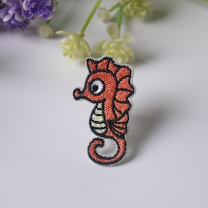 Può includere: Un toppa ricamata da stirare con un cavalluccio marino dei cartoni animati, di colore rosso e bianco con contorno nero.