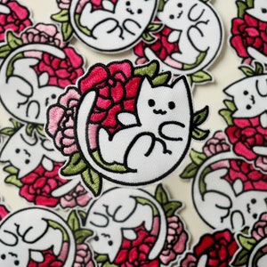 May include: A white embroidered patch of a cat curled up in pink and red peonies with green leaves.