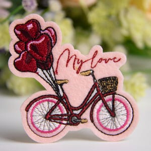 Op de afbeelding: Een roze patch met een rode fiets met een mand en hartvormige ballonnen. De tekst "My love" is in cursief geschreven. De patch heeft een lichtroze rand en is geplaatst tegen een vage bloemenachtergrond.