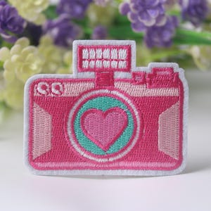 May include: Pink embroidered iron-on patch of a camera with a heart in the viewfinder.
