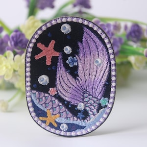 May include: A purple and pink embroidered patch of a mermaid tail with a starfish and bubbles on a dark blue background.