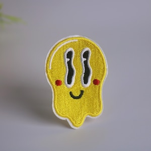 Puede incluir: Parche amarillo bordado para planchar con una cara sonriente y bordes que gotean. La cara tiene dos ojos grandes con pupilas negras y contornos blancos, una boca pequeña y dos mejillas rojas.