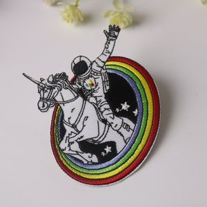 May include: Embroidered patch featuring an astronaut riding a unicorn against a black background with a rainbow border and stars.